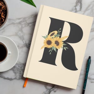May include: A cream-colored journal with a large black letter "R" adorned with yellow sunflowers and green leaves. A cup of coffee and a pen are next to the journal. The journal has an orange ribbon bookmark.