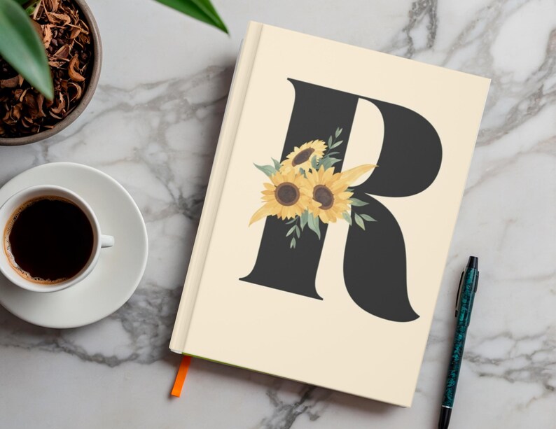 May include: A cream-colored journal with a large black letter "R" adorned with yellow sunflowers and green leaves. A cup of coffee and a pen are next to the journal. The journal has an orange ribbon bookmark.