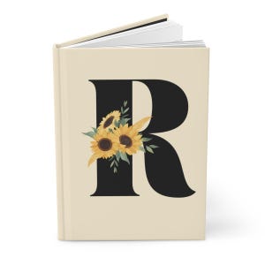 May include: A hardcover journal with a cream-colored cover. A large black letter "R" is centered on the cover, adorned with a watercolor illustration of sunflowers and green leaves. The journal's pages are visible.