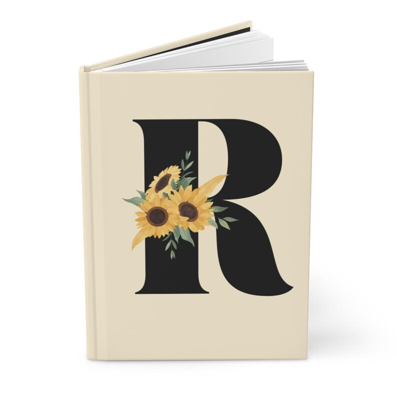 May include: A hardcover journal with a cream-colored cover. A large black letter "R" is centered on the cover, adorned with a watercolor illustration of sunflowers and green leaves. The journal's pages are visible.