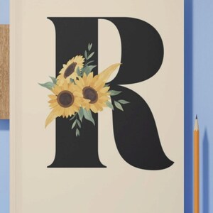 May include: A notebook with a large black letter "R" on the cover, adorned with a watercolor illustration of sunflowers and green leaves. The background is a cream color. A yellow pencil is on the right side.