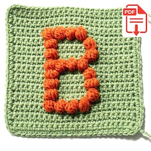 May include: A square crochet piece in light green yarn featuring a large, textured letter B formed by orange bobble stitches. The background is a solid single crochet stitch pattern, and the letter is centered on the square fabric surface.
