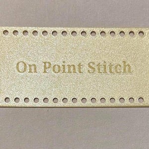 May include: A rectangular, gold-coloured base with evenly spaced holes around the perimeter. The text "On Point Stitch" is engraved in the centre. The base is likely for a craft project, such as a crochet or knitting bag.