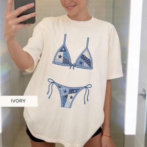 May include: Ivory-colored t-shirt featuring a blue bikini design. The bikini top and bottom are decorated with a patchwork pattern of blue fabrics, including stars and gingham. The word "IVORY" is printed at the bottom left.