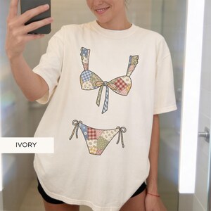 May include: Ivory-colored t-shirt featuring a patchwork bikini design. The bikini top has ruffled straps and a bow, while the bottom has tie sides. The design incorporates various patterns and colors, including blue, red, and floral prints. The word "IVORY" is printed on the bottom left.