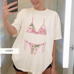 May include: Ivory-colored t-shirt featuring a pink and green bikini design. The bikini top and bottom are decorated with star and patchwork patterns. The word "IVORY" is printed at the bottom of the shirt.