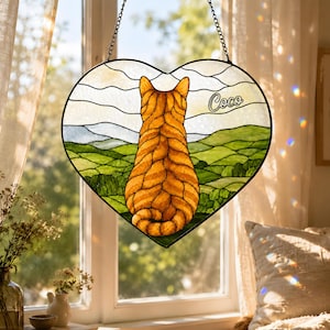 May include: A heart-shaped stained glass suncatcher featuring an orange tabby cat viewed from behind, sitting and looking out over a green rolling landscape under a cloudy sky. The name Coco is written in white script on the glass panel.