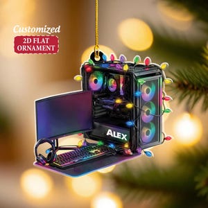 May include: A 2D flat ornament shaped like a computer setup, including a monitor, keyboard, and PC tower. The PC tower has the name "ALEX" on it and is decorated with colorful lights. The ornament is suspended by a gold string.