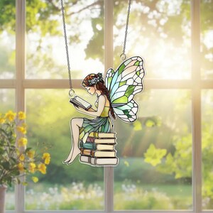 May include: A stained glass fairy reading a book, suspended by a silver chain. The fairy has brown hair, a green and yellow dress, and green and white wings. She is sitting on a stack of books. The background is a window with a view of greenery.