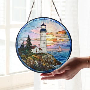 May include: Round stained glass panel depicting a lighthouse on a rocky coast with a sailboat in the distance. The artwork features a colorful sunset sky and is framed with a dark metal chain for hanging. The lighthouse is white with a black top.