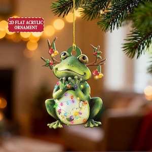 May include: A green frog Christmas ornament with antlers and a string of colorful lights. The ornament is hanging from a gold string and has the text "2D FLAT ACRYLIC ORNAMENT".