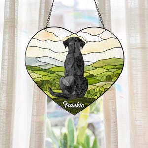 May include: A heart-shaped stained glass suncatcher featuring a black dog sitting with its back to the viewer, overlooking a landscape of green rolling hills and a pale sky. The name Frankie is printed in white script at the bottom of the piece.