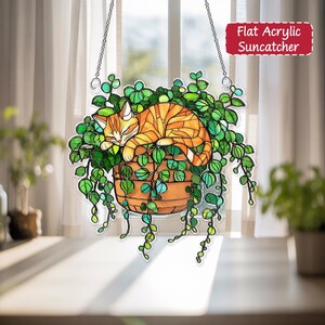 May include: A flat acrylic suncatcher featuring a sleeping orange tabby cat nestled in a hanging basket of green vines. The basket is brown, and the suncatcher hangs from silver chains. The text "Flat Acrylic Suncatcher" is in a red box.