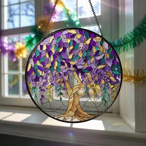 May include: Round stained glass window art featuring a tree with a gold trunk and purple, teal, and gold leaves. The window is framed in black and hangs from a chain. The window is displayed in front of a window.
