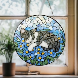 May include: Round stained glass window art featuring a sleeping cat nestled among blue flowers. The cat has gray and white fur. The background is a mosaic of blue and white glass, with a black metal frame and hanging chain.
