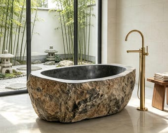 May include: A freestanding bathtub carved from a large, textured natural stone boulder with a smooth, dark interior basin. Beside the tub stands a tall, brushed gold floor-mounted faucet and a small wooden stool holding folded beige towels.