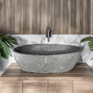 May include: A gray stone vessel sink with a textured surface. The sink is oval-shaped and sits on a wooden cabinet. A black faucet is mounted on the wall above the sink. The background features a wooden panel and a marble backsplash.