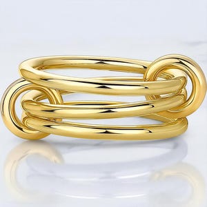 May include: A gold ring with a linked design. The ring has three interconnected bands, creating a unique and modern look. The polished surface reflects light, highlighting the smooth, rounded shape. This piece of jewellery is suitable for everyday wear.