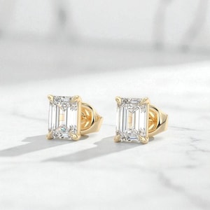 May include: A pair of gold stud earrings featuring emerald-cut, clear gemstones. The earrings are set in a gold bezel setting and are displayed on a white surface. A classic jewellery item.