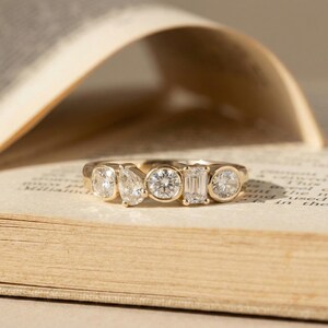 May include: A gold ring featuring five differently shaped diamonds. The diamonds include a square, pear, and round cut, set in a gold band. The ring is displayed on the pages of an open book.