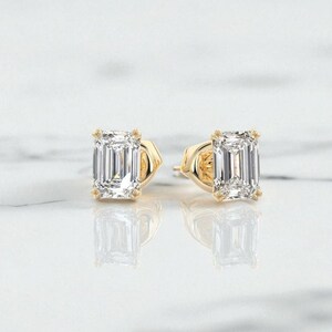 May include: A pair of gold stud earrings with clear, emerald-cut gemstones. The earrings are set in a four-prong setting. The background is a light, neutral colour, highlighting the jewellery. The gemstones reflect light.