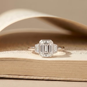 May include: A silver ring with an emerald-cut center stone and two smaller side stones. The ring is set on a light-colored background, possibly a book. The ring's design is classic and elegant, suitable for engagement or special occasions.