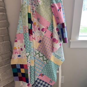 May include: A colorful patchwork quilt draped over a white ladder. The quilt features various patterns, including polka dots, plaid, and floral designs, in shades of pink, blue, green, and yellow. The quilt is handmade and appears soft and cozy.