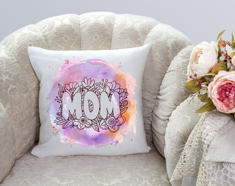 May include: White throw pillow with a watercolor design of flowers and the word "MOM" in a circle. The pillow is on a white armchair with a floral pattern.