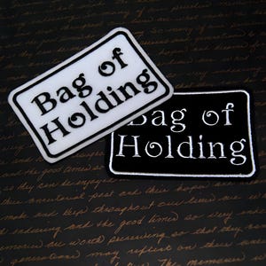 Może przedstawiać: Dwa naszywki do prasowania, jedna biała, a druga czarna, z tekstem "Bag of Holding" w białym, ozdobnym kroju pisma.