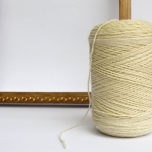 Puede incluir: Una bobina de hilo de color crema enrollada en un pasador de madera. El hilo es liso y tiene un brillo sutil.