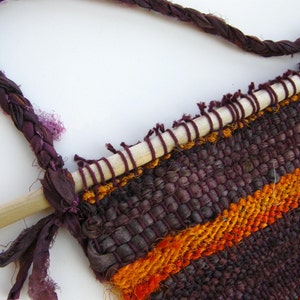 May include: A woven wall hanging with a wooden dowel at the top. The hanging is made of purple, orange, and red yarn and has a fringe at the bottom.