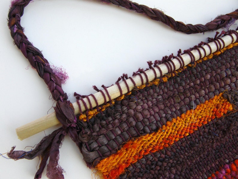 May include: A woven wall hanging with a wooden dowel at the top. The hanging is made of purple, orange, and red yarn and has a fringe at the bottom.