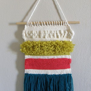 May include: A woven wall hanging with a wooden dowel and white string for hanging. The textile features cream, olive green, red, and teal blue sections. The bottom section has teal blue fringe.