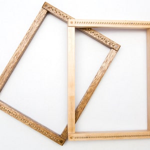 May include: Two wooden weaving looms. One loom is a light brown color, and the other is a darker brown. Both looms are rectangular and have small metal pins along the top and bottom edges, used for holding the weaving threads in place. The looms are set against a white background.