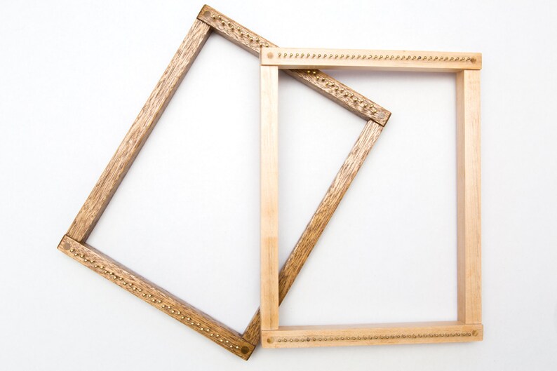 May include: Two wooden weaving looms. One loom is a light brown color, and the other is a darker brown. Both looms are rectangular and have small metal pins along the top and bottom edges, used for holding the weaving threads in place. The looms are set against a white background.