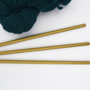 May include: Three gold knitting needles on a white background with a ball of teal yarn.