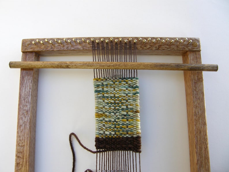 May include: A wooden loom with a brown, green, and yellow woven tapestry on it. The loom has a wooden frame and a wooden bar that is used to move the warp threads.