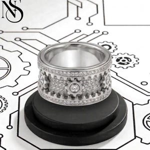 May include: A silver ring with a gear design, encrusted with small, sparkling stones. The ring features a series of gear-shaped cutouts around the band, with a larger stone set in the center of each gear. The ring is displayed on a black stand.