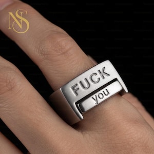 May include: A silver-toned metal ring featuring a rectangular face with the word FUCK engraved in uppercase letters on the top section and the word you engraved in lowercase letters on a separate, slightly recessed lower section of the band.