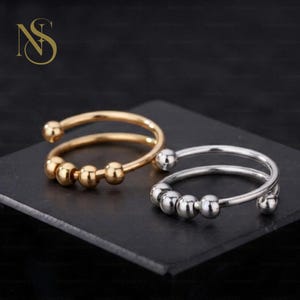 May include: Two open-band rings, one in a gold tone and one in a silver tone, each featuring a smooth, circular wire design with four small, movable spherical beads positioned along the front section of the band for tactile movement.