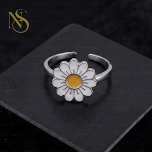 May include: A silver-toned adjustable ring featuring a daisy flower motif. The flower has white petals and a central yellow circular accent. The band is open at the back, allowing for size adjustment. The metal has a smooth, polished finish.