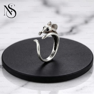 Op de afbeelding: Een zilverkleurige ring met een muisontwerp. De muis heeft een gedetailleerde kop met zwarte ogen en een gebogen staart. De ring wordt gepresenteerd op een zwart cirkelvormig oppervlak.