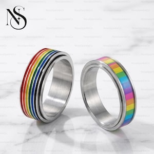 May include: Two stainless steel spinner rings with colorful rainbow designs. One ring features a classic rainbow pattern, while the other has a more pastel, striped design. The rings are displayed on a white surface, with the brand name "NovaSparkleCo" visible.
