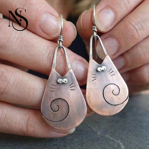 May include: A pair of dangle earrings featuring a copper-toned cat silhouette with a brushed finish. Each earring has a silver-toned wire hook, two small silver-toned dots for eyes, etched whiskers, and a prominent spiral tail design on the body.