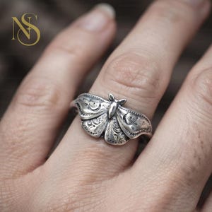 May include: A silver ring featuring a detailed moth design with intricate engraved patterns on the wings. The metal has a polished finish with darkened recessed areas to highlight the texture of the moth's body and wings, worn on a finger.