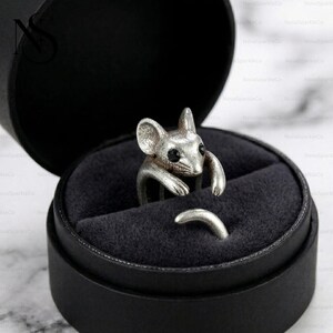 Op de afbeelding: Een zilverkleurige ring in de vorm van een muis, met zwarte ogen, wordt gepresenteerd in een zwarte ringdoos. De armen en de staart van de muis vormen de band van de ring. Een uniek sieraad.