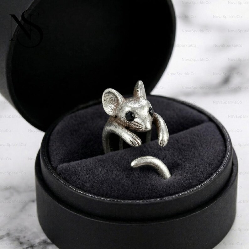 Op de afbeelding: Een zilverkleurige ring in de vorm van een muis, met zwarte ogen, wordt gepresenteerd in een zwarte ringdoos. De armen en de staart van de muis vormen de band van de ring. Een uniek sieraad.