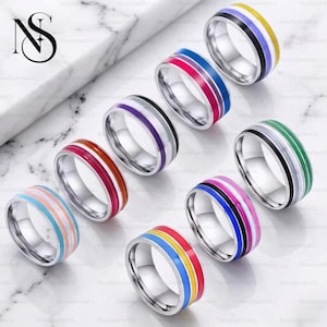 May include: A collection of stainless steel rings with colorful enamel bands. Each ring features a different combination of colors, including red, blue, yellow, pink, and green. The rings are displayed on a marble surface.