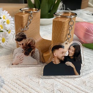 May include: Clear acrylic keychains featuring photographs of couples. Each keychain has a silver-colored chain and ring. The images show couples embracing and smiling. The keychains are displayed on wooden blocks.