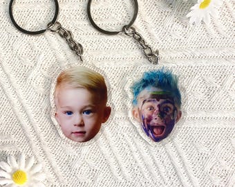 May include: Two clear acrylic keychains with silver metal hardware. One keychain features a photo of a young child with blonde hair and blue eyes. The other keychain has a photo of a child with blue hair and paint on their face.